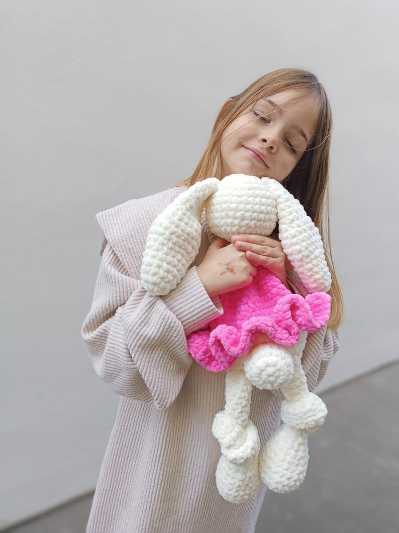 May include: A young girl wearing a beige sweater holds a white crocheted bunny wearing a pink dress. The bunny has large white ears and a fluffy white tail.