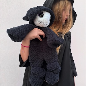 May include: A handmade, dark blue crochet doll in the shape of a grim reaper. The doll has a white face with black stitched features and a black hooded cloak. The doll's arms and legs are also dark blue.
