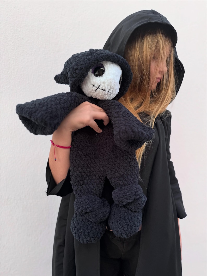 May include: A handmade, dark blue crochet doll in the shape of a grim reaper. The doll has a white face with black stitched features and a black hooded cloak. The doll's arms and legs are also dark blue.