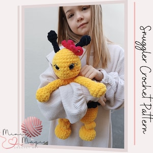 Puede incluir: Un juguete de peluche de abeja tejido a crochet con antenas negras, rayas amarillas y negras y un lazo rosa. La abeja está siendo sostenida por un niño que lleva un suéter blanco.