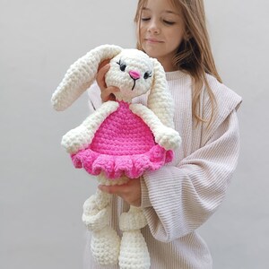 May include: A white crocheted bunny wearing a pink dress with a ruffled hem. The bunny is being held by a young girl.