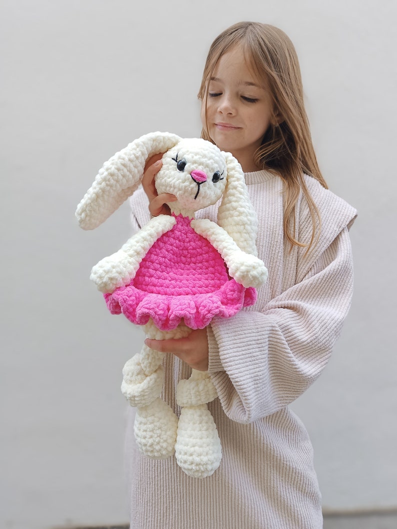May include: A white crocheted bunny wearing a pink dress with a ruffled hem. The bunny is being held by a young girl.