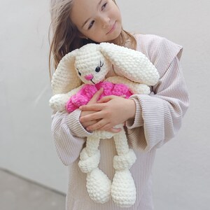 May include: A young girl in a beige sweater holds a white crocheted bunny with pink accents. The bunny has long ears and a pink bow.