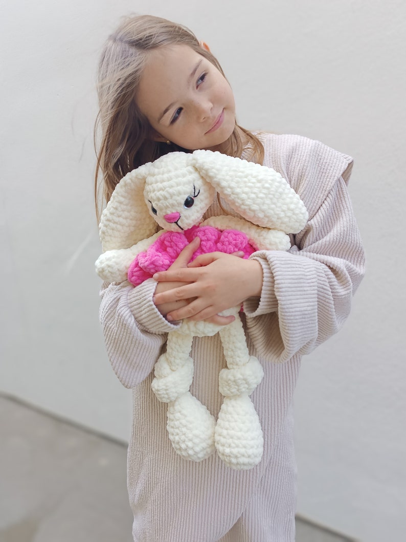 May include: A young girl in a beige sweater holds a white crocheted bunny with pink accents. The bunny has long ears and a pink bow.