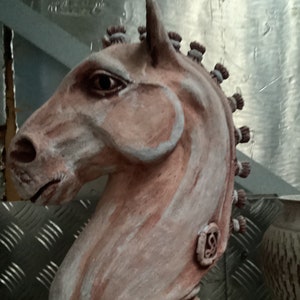 Könnte beinhalten: Eine Keramik-Pferdekopfskulptur mit einem weißen und braunen Farbmuster. Die Skulptur hat eine detaillierte Mähne und einen strukturierten Sockel.