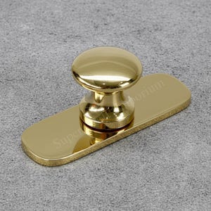 Könnte beinhalten: Ein goldfarbener Schrankknauf mit abgerundeter Oberseite und rechteckiger Basis. Der Knauf ist hochglanzpoliert und reflektiert das Licht, wodurch er glänzend wirkt. Auf der Basis steht "Super Emporium".