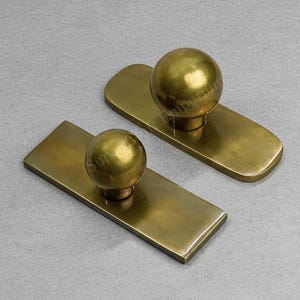 May include: Two brass door pulls, each with a rectangular backplate and a spherical knob. The hardware has a polished finish and a warm, golden hue. The pulls are displayed on a neutral grey surface.
