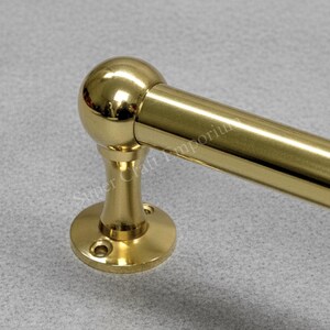 May include: A shiny, gold-coloured towel rail. The rail has a rounded ball at one end and a circular base with screw holes. The finish is reflective, suggesting polished brass or a similar material. The rail is designed for a bathroom.