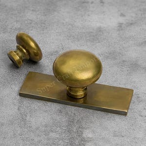 May include: Two antique brass cabinet knobs. One knob is detached, and the other is attached to a rectangular backplate. The knobs and backplate have a warm, golden hue, suggesting an aged or vintage aesthetic. The background is a neutral grey.