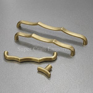 May include: A set of brass-colored cabinet hardware, including two long handles, one shorter handle, and a small knob. The hardware has a curved, flowing design. The text "Super Craft Emporium" is visible.