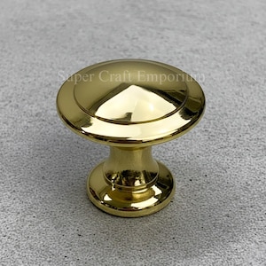 May include: A shiny gold cabinet knob with a round top and a cylindrical base. The knob has a polished finish and the text "Super Craft Emporium" is visible on the top. This hardware is suitable for furniture and home decor projects.