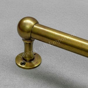 May include: A brass-coloured towel rail with a spherical end and a circular base. The rail is cylindrical and extends to the right. The base has two screw holes. The words "Super Craft Emporium" are printed on the base.
