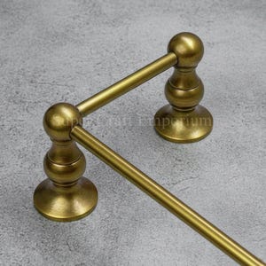 May include: A gold-colored towel rack with a classic design. The rack features two horizontal bars supported by decorative posts with rounded finials and circular bases. The metal has a slightly aged appearance.