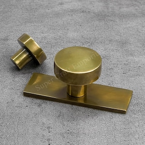 Könnte beinhalten: Zwei Messing-Schrankknöpfe. Ein Knopf hat eine zylindrische Form mit rechteckiger Basis, der andere ist ein kleinerer Zylinder. Die Knöpfe haben eine warme, metallische Farbe und sind vor einem grauen Hintergrund platziert.