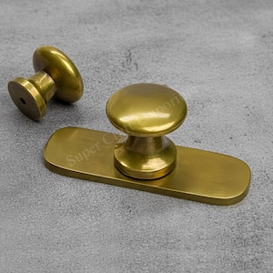 Peut inclure: Deux boutons de meuble en laiton. L'un des boutons est détaché, montrant le trou de vis. L'autre bouton est fixé à une plaque arrière rectangulaire en laiton. Les boutons et la plaque arrière ont une finition polie. Le fond est gris moucheté.