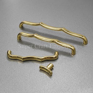 May include: Four brass cabinet hardware pieces. Two curved handles, one smaller handle, and a knob. The hardware is gold-colored and has a shiny finish. The pieces are arranged on a gray surface.
