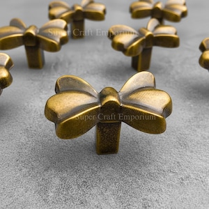 May include: Antique brass bow-shaped cabinet knobs. The knobs have a bow design with a rectangular base. The text "Super Craft Emporium" is visible on some of the knobs. These decorative knobs are suitable for furniture.