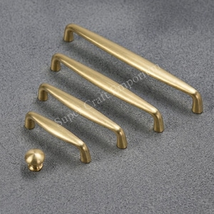 May include: A set of brass cabinet handles and a knob, in varying sizes. The hardware has a brushed finish and a warm, golden colour. The items are arranged diagonally on a grey surface. The knob is round.