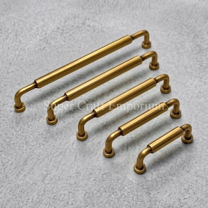 May include: Five brass-toned metal cabinet pulls arranged in a diagonal line from longest to shortest. Each handle features a cylindrical bar with rounded ends and two perpendicular mounting posts that connect to the base of the hardware.