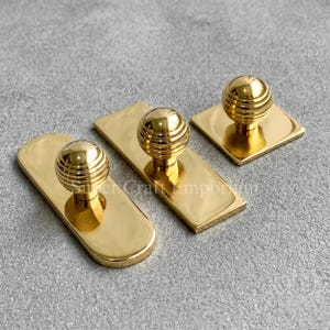 Könnte beinhalten: Drei goldfarbene Schrankknöpfe mit geripptem Kugeldesign. Zwei Knöpfe sind auf rechteckigen Platten montiert, einer auf einer quadratischen Platte. Die Hardware wird auf einer grauen Oberfläche präsentiert.