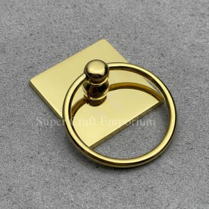 May include: A gold-coloured metal ring pull with a square base. The ring is circular and attached to a small, square plate. The text "Super Craft Emporium" is visible on the base. The hardware is likely for furniture or crafts.
