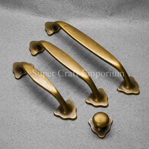 Könnte beinhalten: Ein Satz antiker Messing-Schrankbeschläge, darunter drei Griffleisten und ein Knopf. Die Griffe haben ein geschwungenes Design mit dekorativen blütenförmigen Basen. Der Knopf hat eine passende Blumenbasis. Der Text "Super Craft Emporium" ist sichtbar.