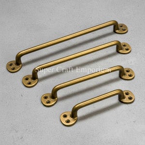 Könnte beinhalten: Vier antike Messing-Schrankgriffe in verschiedenen Größen. Jeder Griff hat einen gebogenen Griff und runde Montageplatten mit drei Schraubenlöchern. Die Griffe sind diagonal auf einer grauen Oberfläche angeordnet. Der Text "Super Craft Emporium" ist sichtbar.