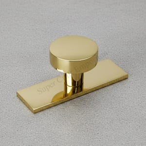 Könnte beinhalten: Goldfarbener Schrankknopf mit rechteckiger Basis und rundem Oberteil. Der Knopf hat eine polierte Oberfläche und befindet sich vor einem grauen Hintergrund. Der Text "Super C Emporium" ist auf der Basis sichtbar.