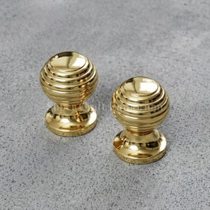 May include: Two polished gold-tone metal cabinet knobs with a spherical, ribbed design. Each knob features a smooth, rounded top and a flared circular base, showcasing a reflective, metallic finish with horizontal grooves circling the sphere.