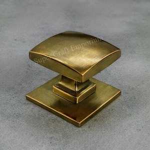 May include: A square brass cabinet knob with a curved top and a square base. The knob has a vintage, antique appearance. The words "Super Craft Emporium" are printed on the top of the knob.
