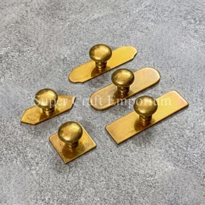 May include: A collection of five brass cabinet knobs, each featuring a round handle attached to a flat metal backplate. The backplates vary in shape, including rectangular, square, and hexagonal designs, all with a polished gold-toned finish.