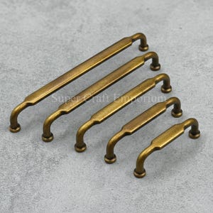 May include: Five antique brass cabinet pulls of varying lengths are arranged diagonally. Each pull has a rectangular centre and curved ends. The pulls are a warm, aged brass colour, suggesting a vintage aesthetic. The text "Supercraft Emporium" is visible.
