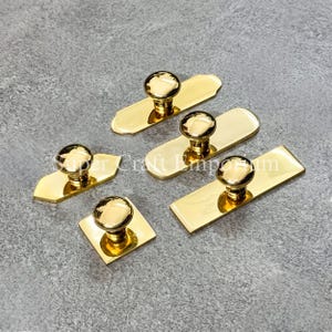 May include: Five polished gold-tone metal cabinet knobs, each featuring a round handle attached to a flat base plate. The base plates vary in shape, including rectangular, square, and rounded-end designs. The text Super Craft Emporium is visible.