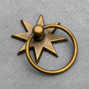 Könnte beinhalten: Antiker Schubladengriff aus Messing mit Sternenmuster. Der Griff hat eine siebenstrahlige Sternbasis und einen kreisförmigen Griff. Die Beschläge sind wahrscheinlich für Möbel oder Schränke und verleihen eine Vintage-Ästhetik.