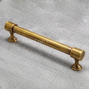 May include: A cylindrical cabinet pull handle with a brushed antique brass finish. The hardware features a long, straight bar supported by two circular pedestals at each end, providing a sturdy grip for opening drawers or cabinet doors.