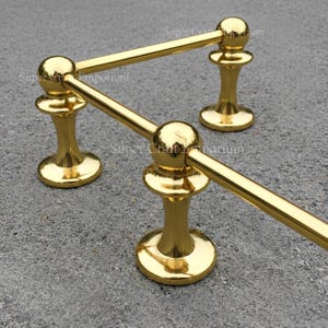 May include: A gold-colored decorative railing system with three vertical supports and horizontal bars. Each support has a rounded base and a spherical top, connected by straight bars. The design is symmetrical and shiny.