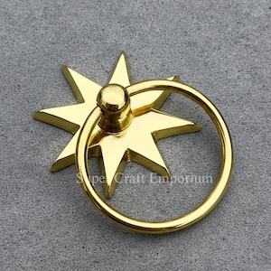 May include: A gold-coloured star-shaped cabinet pull with a circular handle. The hardware features a starburst design with eight points. The pull is made of metal and is likely used for drawers or cabinets.