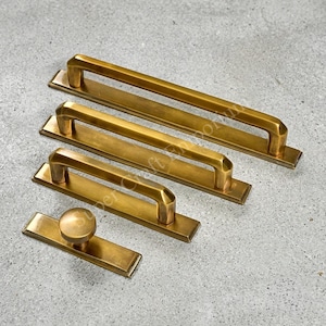May include: A collection of four brass-toned hardware pieces including three rectangular drawer pulls of varying lengths and one circular knob, each mounted on a flat rectangular base plate with a smooth, metallic finish and bevelled edges.