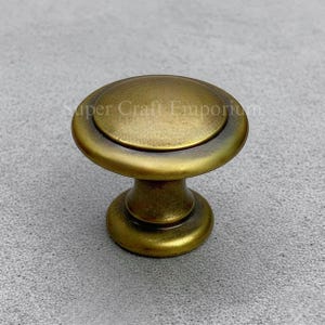 May include: A bronze-colored cabinet knob with a rounded top and base. The knob has a classic design and is likely made of metal. The text "Super Craft Emporium" is visible on the knob.