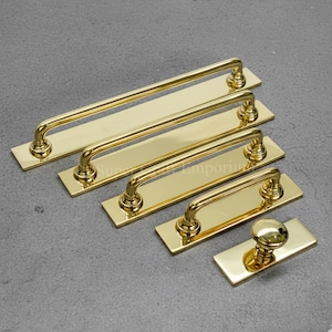 May include: A selection of polished brass door handles and a knob, displayed on a grey surface. The handles are of varying lengths, with a curved bar design fixed to rectangular backplates. The knob has a rounded top and is also on a rectangular backplate.