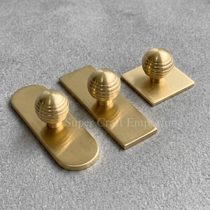 May include: Three brass cabinet pulls with a gold finish. The pulls have a ribbed, spherical knob design. One pull is square, and the other two are rectangular with rounded edges. The pulls are arranged on a grey surface.
