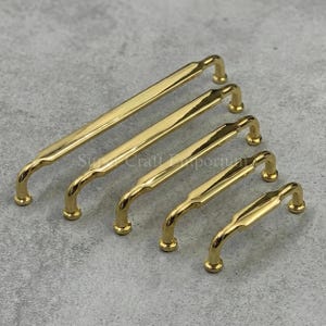 Könnte beinhalten: Fünf goldfarbene Schrankgriffe in verschiedenen Größen. Jeder Griff hat einen gebogenen Griff und abgerundete Enden. Die Griffe sind diagonal auf einer grauen Oberfläche angeordnet und zeigen ihre polierte Oberfläche und ihr Design.