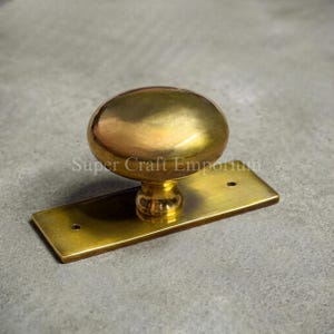 May include: A polished brass door knob featuring a smooth, rounded oval shape mounted on a rectangular brass backplate with two visible screw holes. The metal surface has a reflective, golden finish. The text Super Craft Emporium is visible.