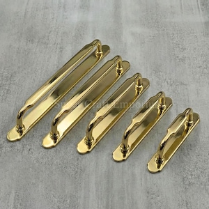 May include: Five brass-coloured door pulls of different sizes. Each pull has a rectangular backplate and curved handles. The pulls are arranged diagonally, highlighting their polished finish and design. Measurements in cm.