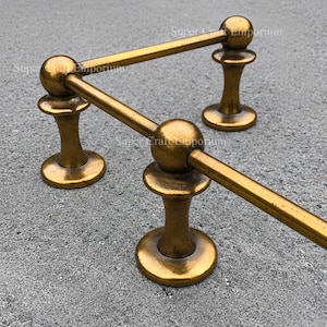 May include: A gold-colored, decorative towel rack with a unique design. The rack features three vertical supports with a round base and a ball-shaped top, connected by horizontal bars. The metal has a slightly aged appearance.