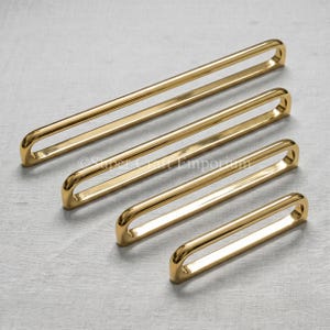 Peut inclure: Quatre poignées de placard en métal doré poli, disposées en diagonale de la plus longue à la plus courte. Chaque poignée présente un design en boucle allongée et continue avec des bords arrondis et une finition lisse et réfléchissante. Le texte Super Craft Emporium est visible.