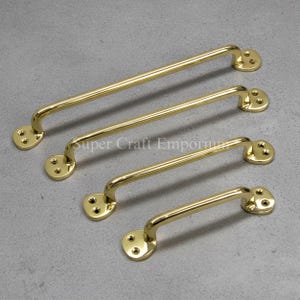 Könnte beinhalten: Vier polierte Messing-Schrankgriffe in verschiedenen Größen. Jeder Griff hat einen gebogenen Griff und zwei kreisförmige Montageplatten mit drei Schraubenlöchern. Die Griffe sind diagonal auf einer grauen Oberfläche angeordnet.