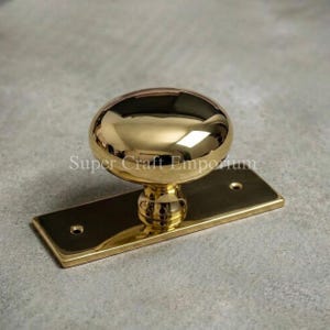 May include: A polished brass door knob with a smooth, rounded, reflective surface attached to a rectangular brass backplate. The backplate features two circular screw holes, one at each end, and the text Super Craft Emporium is visible.