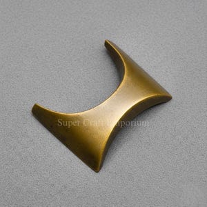 Könnte beinhalten: Ein bronzefarbener, dekorativer Möbelgriff mit geschwungenem, eckigem Design. Der Griff hat eine dreieckige Form mit abgerundeten Kanten und einer leicht strukturierten Oberfläche. Der Text "Super Craft Emporium" ist auf dem Griff aufgedruckt.