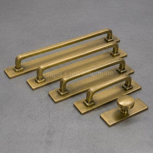Peut inclure: Un ensemble de cinq pièces de quincaillerie en laiton antique comprenant quatre poignées de tiroir rectangulaires de longueurs variables et un bouton circulaire, montés sur des plaques rectangulaires avec une finition brossée.